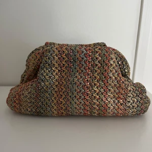 Flerfärgad stickad handväska - Säljer en unik flerfärgad stickad handväska med ett  mönster i guld, rött och grönt. Väskan har en rymlig insida med svart foder och stängs med en praktisk dragkedja. Perfekt för att ge din outfit en färgglad touch!