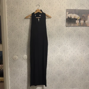 Svart långklänning med öppen rygg - Elegant svart långklänning med en stilren design och öppen rygg. Perfekt för festliga tillfällen. Klänningen har en v-ringad hals och är ärmlös,  ger en sofistikerad look.