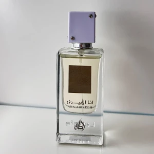Ana Abiyedh parfym 60 ml - Elegant parfym från Lattafa i en stilren, rektangulär glasflaska med en ljuslila kork. Flaskan har en dekorativ etikett med arabisk text och ett intrikat mönster. Doften är sofistikerad och passar perfekt för den som söker något unikt.