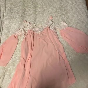 Rosa offshoulder klänning från Parisia - Säljer en söt rosa offshoulder klänning från Parisia med vackra spetsdetaljer vid axlarna. Klänningen har en luftig passform och är perfekt för varma dagar. Den är tillverkad i ett lätt och bekvämt material. Kan finnas lite trådar som hänger löst, men är inget som störs