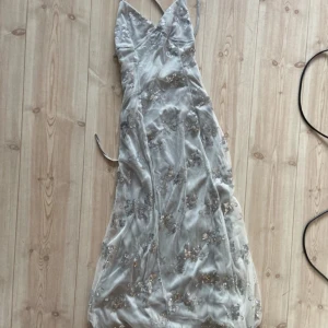 Glittrig långklänning med paljetter - Elegant långklänning i silver färg med tunna axelband,  v-ringad design och öppen i ryggen. Klänningen är dekorerad med glittriga paljetter i blommönster och har en dragkedja på baksidan/ryggen. Perfekt för festliga tillfällen och bal. Storlek M (oanvänd) 