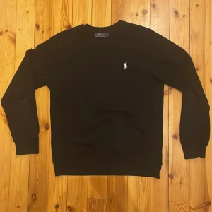 Svart tröja från Polo Ralph Lauren - Snygg svart tröja från Polo Ralph Lauren med det klassiska vita logotypbroderiet på bröstet. Tröjan har långa ärmar och en rund halsringning. Perfekt för en stilren och avslappnad look.