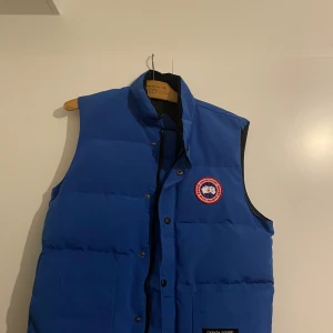 Blå dunväst från Canada Goose - Säljer en blå dunväst från Canada Goose med deras ikoniska logga på bröstet. Västen har en hög krage och knappar framtill. Perfekt för kyliga dagar när du vill hålla värmen utan att kompromissa med stilen.