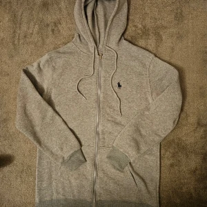 Grå hoodie från Ralph Lauren - Säljer en stilren grå hoodie från Ralph Lauren med dragkedja och klassisk logga på bröstet. Tröjan har långa ärmar och en praktisk huva med dragsko. Perfekt för en avslappnad look. Inte äkta