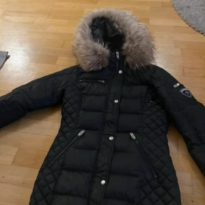 Svart dunjacka med päls från Rockandblue - Snygg svart dunjacka från Rockandblue med quiltad design och avtagbar päls på huvan. Jackan har dragkedja och knappar framtill samt flera praktiska fickor med dragkedjor. Perfekt för kalla dagar!