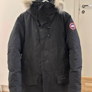 Svart parkajacka från Canada Goose - Säljer en svart parkajacka från Canada Goose med pälsdetaljer på huvan. Jackan har en dragkedja och knappar framtill samt långa ärmar. Perfekt för kalla vinterdagar.