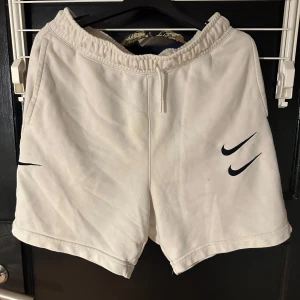 Vita shorts från Nike - Säljer ett par vita shorts från Nike med elastisk midja och snörning. De har en stilren design med svarta Nike-loggor på framsidan och en bakficka med textdetaljer. Perfekta för en avslappnad stil.