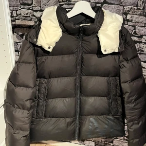 Svart dunjacka från Moncler - Snygg svart dunjacka från Moncler med vit insida på huvan. Jackan har en dragkedja framtill och är perfekt för kyliga dagar. Den har en klassisk pufferdesign som ger både stil och värme. Vid frågor eller funderingar så är de bara o slå en pling👌