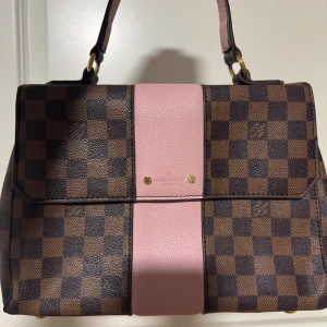 Rutig handväska med rosa detaljer - Snygg handväska från Louis Vuitton i brunt och rosa. Väskan har ett klassiskt rutmönster och en bred rosa remsa i mitten. Den har två handtag och guldiga detaljer. Perfekt för att ge din outfit en lyxig touch.