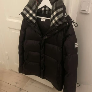 Svart dunjacka från Burberry - Säljer denna snygga dunjacka från Burberry eftersom den är lite för liten för mig. Jackan har en dragkedja och knappar framtill samt en hög krage för extra värme. Perfekt för kyliga dagar. Den är i mycket bra skick då jag bara använt den ungefär 10 gånger sen jag köpte den.