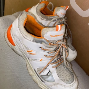 Vita och orange sneakers med snörning - Säljer ett par vita sneakers med orange detaljer och snörning. Skorna har en sportig design med texten 'GodblessAir' på sidan och tungan. Perfekta för en casual look. Den behöver tvättas då den har legat på källaren