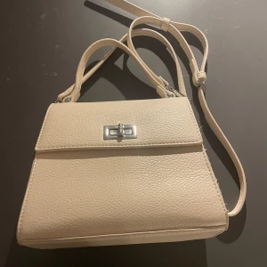 Beige axelväska med silverdetalj - Snygg beige axelväska i skinn med en elegant silverdetalj på framsidan. Väskan har en justerbar axelrem och en stilren design som passar perfekt för alla tillfällen.