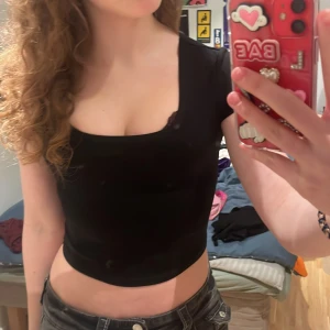 Svart croppad topp från Bik Bok - Säljer en stilren svart croppad topp från Bik Bok. Toppen har en djup rundad hals och korta ärmar, perfekt för en trendig look. Passar bra till både jeans och kjol.