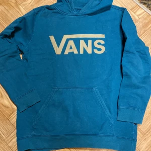 Blå hoodie från Vans - Säljer en snygg blå hoodie från Vans med deras ikoniska logga i vitt på bröstet. Tröjan har en klassisk känguruficka och ribbade muddar vid ärmslut och nederkant. Perfekt för en avslappnad stil. Hoodien är använd men är i gott skick   Jag tror att det är barn storlek xl men den passar tjej xs/s   Färgen är blå/grön 
