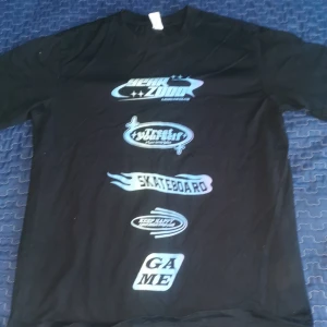Svart t-shirt med skateboardtryck - Cool svart t-shirt med flera tryck i blått och vitt, inklusive skateboardmotiv och text som 'GAME'. Perfekt för en avslappnad stil.