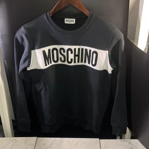 Svart sweatshirt från Moschino - Snygg svart sweatshirt från Moschino med stort vitt tryck på bröstet. Tröjan har rund hals och långa ärmar, perfekt för en stilren look. Passar både till jeans och joggers. Det finns fläckar som syns på bild 4 och 5, annars fint skick. Hör av er vid minsta fundering🙌