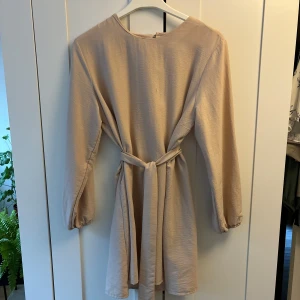Beige långblus med knytband - Långärmad blus från Modanisa med ett avtagbart bälte i samma tyg. Kan kanske användas som en kortare klänning. Storlek 44. Säljer då den är för stor och jag inte använder den längre. Materialet är skönt och luftigt. 100% polyester.