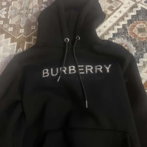 Svart hoodie från Burberry - Säljer en stilren svart hoodie från Burberry med logotypen broderad på framsidan. Hoodien har en klassisk design med dragsko i huvan och en stor ficka framtill. Perfekt för en avslappnad och trendig look.