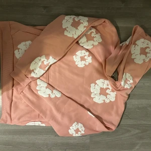 Rosa hoodie från Denim Tears - Säljer en rosa hoodie från Denim Tears med vita blommönster. Tröjan har långa ärmar och en klassisk huva. Perfekt för en avslappnad stil.