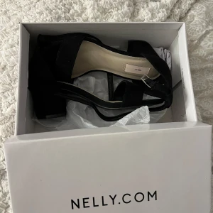 Svarta klackskor från Nelly - Säljer ett par grymt snygga svarta klackskor från Nelly. De har en bred rem över tårna och en justerbar rem runt ankeln. Skorna har en bekväm blockklack. Skorna är i nyskick och har kvar orginalkartongen som skorna kom i🩷