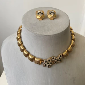 Guldhalsband och örhängen med blommotiv - Vintage elegant set med guldhalsband och matchande örhängen. Halsbandet har en unik design med stora, guldfärgade länkar och ett vackert blommotiv i mitten dekorerat med små stenar. Örhängena är fyrkantiga med samma blommotiv och stenar. Okänd metall