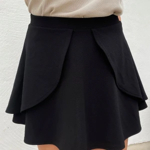 Svart kortkjol med volangdetaljer - Säljer en superfin svart kortkjol med volangdetaljer. Kjolen har en skön passform och är perfekt för både vardag och fest. Den är gjord i ett mjukt material som faller snyggt. 