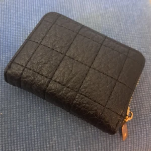 Svart quiltad plånbok med dragkedja - Säljer en snygg och praktisk svart plånbok med quiltat mönster. Den har en dragkedja i guld och flera fack inuti för kort och kontanter. Perfekt för att hålla ordning på dina grejer! oanvänd