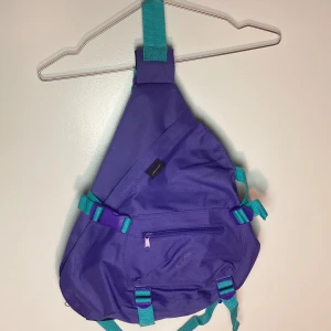 Lila crossbody crossbody ryggsäck med blå detaljer - Lila crosbody ryggsäck med blå spänndetaljer. Många fickor och fack. Mellanstor i sin storlek. Går att spänna om med kardborr efter storlek