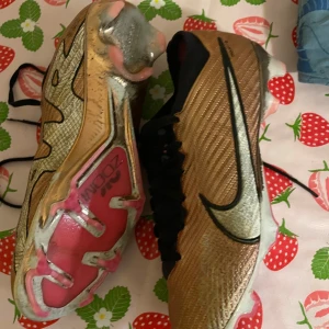 Guldiga fotbollsskor med rosa detaljer - Säljer ett par coola guldiga fotbollsskor från Nike med rosa detaljer på sulan. Skorna har en snygg svart snörning och är perfekta för fotbollsträningen. De är i bra skick och redo för nya äventyr på planen!