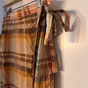 Rutig omlottkjol i höstfärger - Säljer en superfin rutig omlottkjol i härliga höstfärger som orange, brun och beige. Kjolen har ett knytband i sidan för en justerbar passform. Perfekt för höstpromenader eller en mysig fika på stan. Materialet känns lätt och bekvämt.