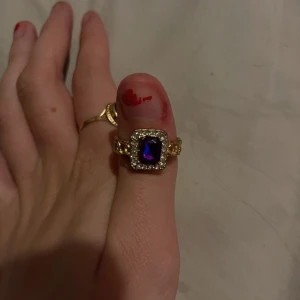 Guldring med lila sten - Säljer en elegant guldring med en stor lila sten i mitten, omgiven av små gnistrande stenar. Perfekt för att ge en touch av glamour till vilken outfit som helst. Ringen har en klassisk och tidlös design.