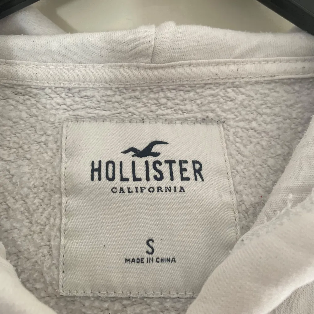 Säljer en vit hoodie från Hollister i storlek S. Den har en dragkedja framtill och en stor logga på ärmen. Perfekt för en avslappnad stil och passar bra till både jeans och mjukisbyxor. Den är i bra skick och supermjuk! 🕊️. Tröjor & Koftor.