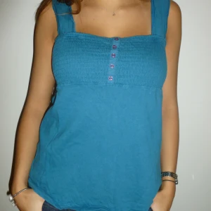 Blå topp - Somrig blå topp med knappar framtill. Perfekt för varma dagar och kan enkelt matchas med jeans eller shorts. Toppen har en smockad detalj vid bysten som ger en fin passform.