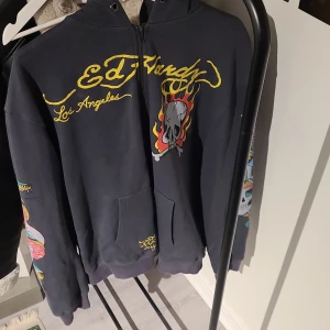 Svart hoodie från Ed Hardy - Säljer en svart hoodie från Ed Hardy med coola tryck av dödskallar och eldflammor. Tröjan har dragkedja och långa ärmar med färgglada detaljer på ärmarna och ryggen. Perfekt för dig som gillar en edgy stil!