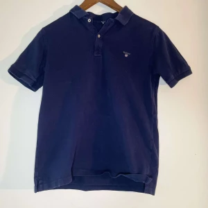  pikétröja från GANT - Säljer en klassisk mörkblå pikétröja från GANT. Tröjan har korta ärmar och en stilren krage med knappar framtill. Perfekt för en avslappnad men ändå stilfull look.