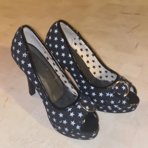 Svarta pumps med stjärnmönster - Snygga svarta pumps med vita stjärnor och öppen tå. Skorna har en hög klack och en dekorativ ringdetalj framtill. Perfekta för att ge en unik touch till din outfit.