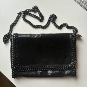 Svart axelväska med kedjedetaljer - Svart axelväska med kedjedetaljer runt kanterna och en matchande kedjerem. Tillverkad i ett glansigt material.