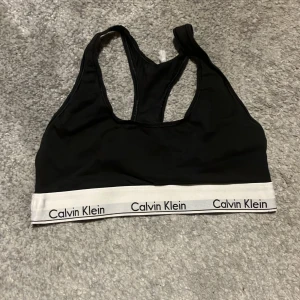 Svart sporttopp från Calvin Klein - Säljer en svart sporttopp från Calvin Klein med vit resårkant och logga. Toppen har en racerback-design och är perfekt för träning. Bekväm och snygg. Jätte bekväm går både att sova me och bara ha på sig eller träna med! De är och ser använd ut men är skön och är inte sönder. Pris kan alltid diskuteras och skriv till mig för övriga frågor eller bilder!💗