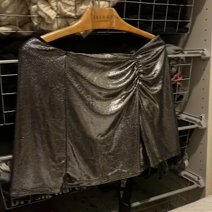 Glittrig silverkjol - Snygg glittrig silverkjol med en stilren design. Kjolen har en smickrande drapering framtill och en kort längd som ger en festlig look. Perfekt för att sticka ut på nästa event!