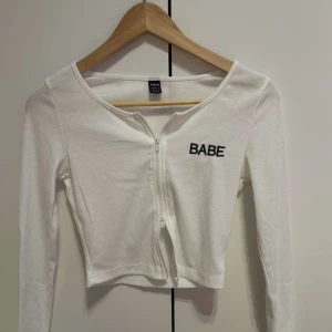 Vit ribbad bolerotopp med dragkedja - Säljer en vit ribbad bolerotopp från Zara med texten 'Babe' broderad på bröstet. Toppen har en dragkedja framtill och långa ärmar. Perfekt för en trendig look!