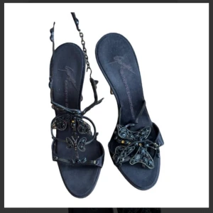 Svarta sandaler med fjärilsdetaljer - Eleganta svarta sandaler med dekorativa fjärilsdetaljer. Sandalerna har en smal remdesign och en liten klack, vilket ger en stilren look. Perfekta för sommarens alla tillfällen.