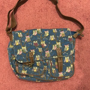 Blå axelväska med ugglemönster - Charmig blå axelväska med färgglatt ugglemönster. Väskan har bruna detaljer och justerbar axelrem. Perfekt för en unik och lekfull stil.