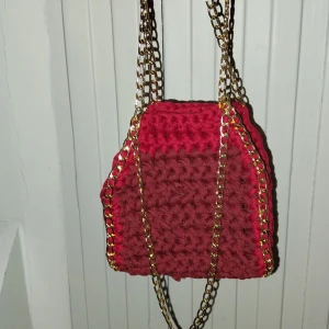 Vinröd virkad väska med kedja - Snygg vinröd virkad väska med guldiga kedjedetaljer. Väskan har en unik textur och är perfekt för att ge din outfit en färgklick. Den har en praktisk storlek och kan bäras i handen.