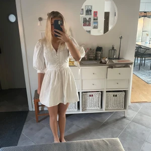 Vit kortklänning med v-ringning - Säljer en söt vit kortklänning med v-ringning och trekvartsärmar. Klänningen har fina detaljer med knappar framtill och en lätt volang vid ärmsluten. Perfekt för sommarens alla tillfällen! 🌸