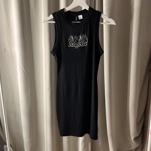 Svart ärmlös klänning med tryck - Snygg svart ärmlös klänning med texten 'Angelic' och ett coolt tryck på bröstet. Perfekt för en avslappnad stil med en edgy touch. Klänningen har en enkel och stilren design som gör den lätt att matcha med olika accessoarer.