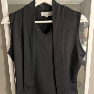 Svart ärmlös topp från Amalia - Snygg svart ärmlös topp från Amalia med en elegant knytning i nacken. Perfekt för en stilren look. Toppen har en v-ringad design och är tillverkad i ett mjukt material.