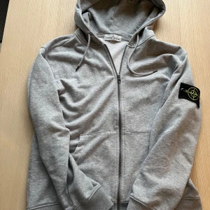 Grå hoodie från Stone Island - Säljer en stilren grå hoodie från Stone Island med dragkedja och klassisk logga på ärmen i storlek L men skulle säga att den är som S/M. Den har luva och fickor framtill. Perfekt för dig som gillar en avslappnad stil.