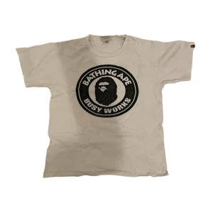 Vit A Bathing Ape t-shirt - Säljer en vit t-shirt från A Bathing Ape med det ikoniska 'Busy Works' trycket på framsidan. T-shirten har en rund hals och korta ärmar. Perfekt för dig som gillar streetwear och vill ha något unikt i garderoben.