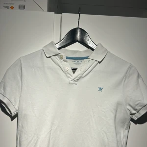 Vit pikétröja från Hackett - Säljer en stilren vit pikétröja från Hackett med broderad logga på bröstet. Tröjan har korta ärmar och klassisk krage med knappar. Perfekt för en avslappnad men ändå snygg look.