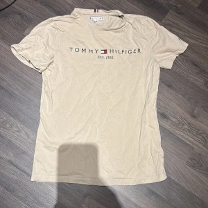 Beige t-shirt från Tommy Hilfiger - Säljer en beige t-shirt från Tommy Hilfiger med tryck på framsidan. Storlek 164